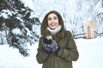 Obraz premium young beautiful brunette woman in a warm jacket with hoodie and warm mittens is walking outdoors in winter. a woman holding a paper glass with hot tea or coffee. Enjoys a walk on a frosty day