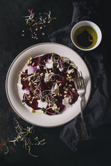 Obraz na płótnie Canvas Vegetarian beetroot carpaccio salads with olive oil, goat cheese, pine nuts and sprouts in white plate on textile napkin over dark rusty metal background. Dark rustic style, healthy eating