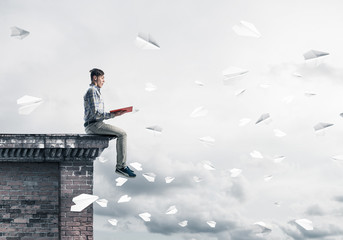 Handsome student guy on roof edge reading book and paper planes 