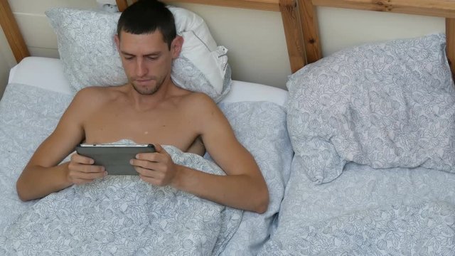Young man is typing something on a lepton or tablet lying on the bed stretching and yawning