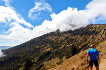 Fototapeta na wymiar Sporty guy traveler in a blue t-shirt on a background of a beautiful sky with clouds in the mountains. Trekking to Rinjani Volcano. Indonesia. Lombok Island.