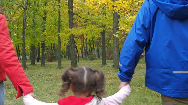 Parents with the child walk in the autumn park. A little girl is holding her parents by the hands.