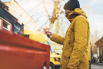 Fototapeta na wymiar Hipster with yellow backpack, jacket, cap, coffee of thermo cup holding in the hands smartphone, freelance using gadget mobile in autumn street outdoor, tourist man typing message, online process