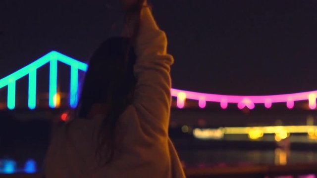 Slow motion. a young blonde girl with long curly hair dances in the night gown on the background of glowing fanfare. bright night illumination and a luminous bridge. The professional dancer moves