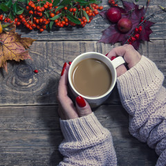 Female hands holding cup of coffee on autumn wooden background. Old book, autumn leaves and dried rowan on background. Concept cozy coffee cup. Top view