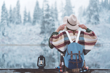 Young traveling woman sitting with kerosene lamp in front of amazing frozen landscape. Wearing hat, poncho and backpack. Winter is coming, first snowfall. Wanderlust and boho style