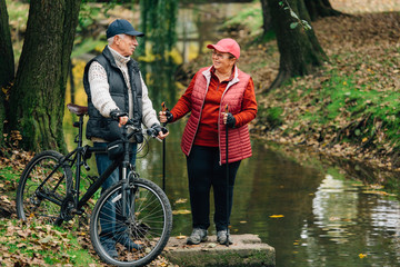 Pretty senior sport couple standing  in colorful autumn park near river. Mature woman with nordic...