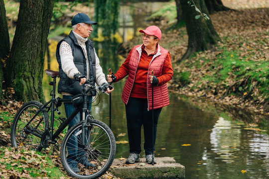 Pretty senior sport couple standing  in colorful autumn park near river. Mature woman with nordic walking poles and old man with bicycle resting outdoors.