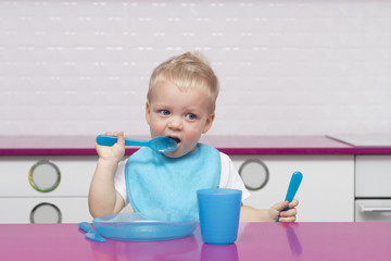 Portrait Of cute blonde toddler in a blue bib with fork and knife in his hands In High Chair in the modern kitchen ready to eat