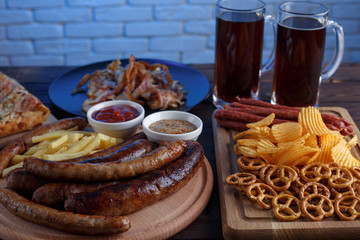 Dark beer and appetizing beer snacks set. Close up of two mugs of stout and grilled sausages with french fries, salami, chips, pretzel crackers and sauces on the table. Oktoberfest food, pub concept