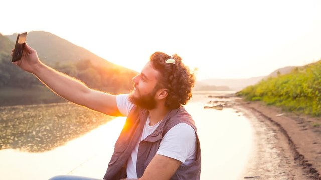 A stylish young fisherman sits in a wooden boat and doing a fun Selfie against the backdrop of the sunset and river