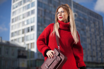 Fashionable girl in a red sweater against the backdrop of city bustle. In glasses and with a brown...