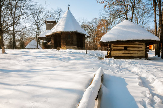 A small wooden church and cottage covered in snow at the Village Museum, an open-air ethnographic museum located in Bucharest, Romania.