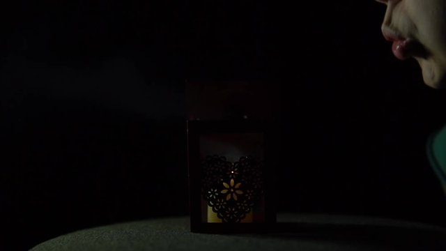 Young woman blowing on a burning candle in a metal lantern with an openwork heart on a black background. The flame goes out, the smoke remains.
