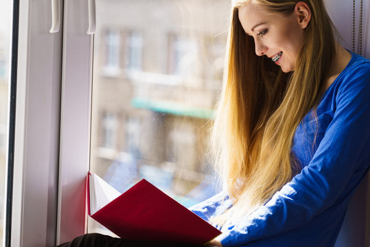 Woman sitting on window sill reading book at home