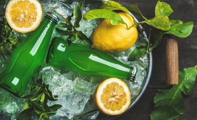 Bottles of green lemonade on chipped ice in metal tray with fresh lemons and mint over dark wooden background, top view - obrazy, fototapety, plakaty