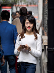 Front view of a happy Chinese girl walking and using a smart phone in crowded street.