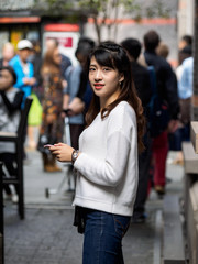 Front view of a happy Chinese girl walking and using a smart phone in crowded street.