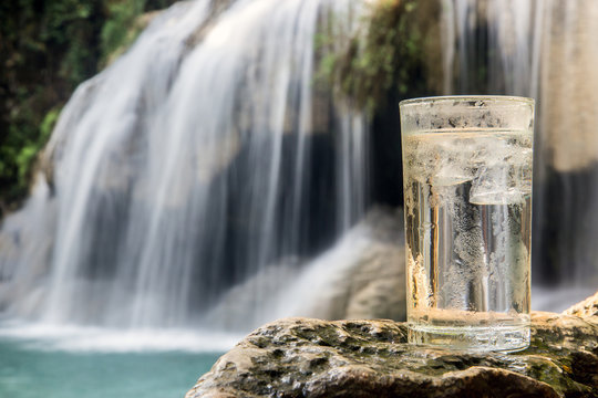Dewy glass full of water with ice cube stands on a rock near a waterfall in the forest. A refreshing drink from nature.