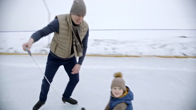 Handheld tracking of young man and woman skating on outdoor ice rink and pulling sled with cute little boy on warm winter day