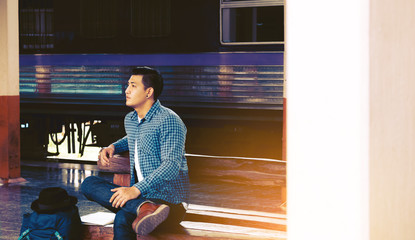 asian hipster young man sitting at train station with awaiting the train's arrival.