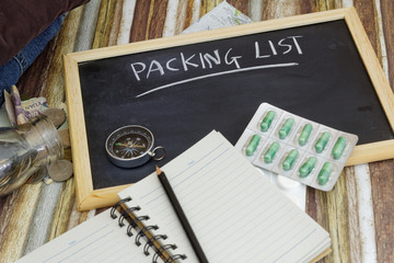 Packing list on blackboard with money spilling out of glass jar, compass,medicine blister pack, stack of clothes,pencil,map and blank notepad