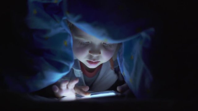 Little child, with blue eyes, watching a digital tablet under the bed sheets