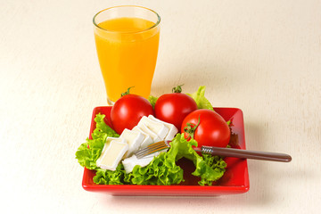 Tomatoes and cheese slices lie on a sheet of fresh salad in a red plate. A glass of orange juice stands side by side on the table.