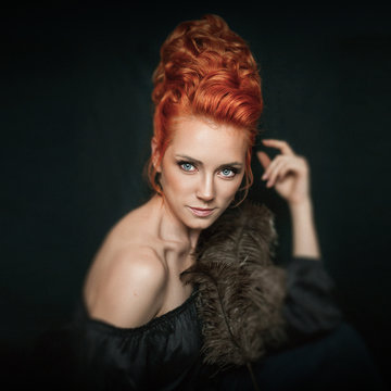 Portrait of redhead woman with blue eyes wearing black dress on dark background. Girl doesn't look into the camera. Female is posing with bird feather.