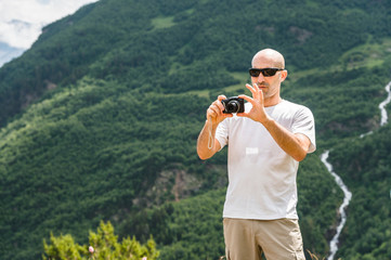 A bald tourist in sunglasses at a halt takes a picture on his camera. Against the backdrop of green mountains and a river