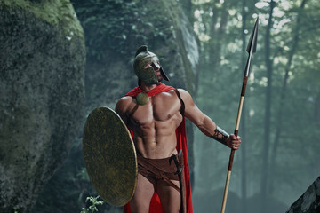 Spartan warrior in a battledress walking in the woods with his weapon.