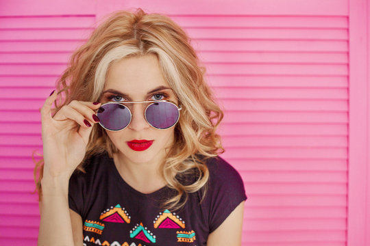 Fun and colorful.  Close up portrait of Young pretty happy blonde woman in shorts and sunglasses posing against pink background. Beauty and fashion concept.