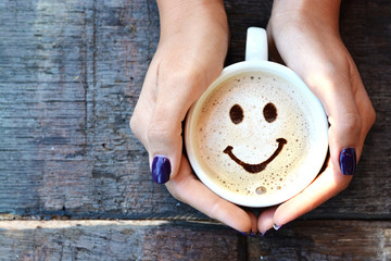 Woman hands holding one cappuccino cup with optimistic face on wooden table - obrazy, fototapety, plakaty
