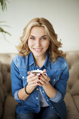 Starting new day with coffee. Beautiful cheerful young blonde woman holding coffee cup with smile while sitting on the sofa at home.