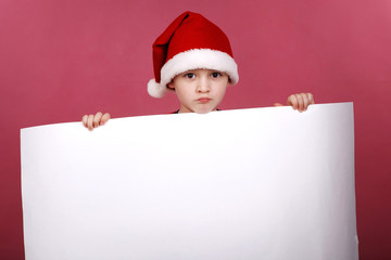 Portrait of little boy in the hat of Santa Claus on a red background , holding a white sheet of paper