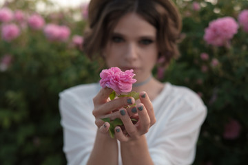 Beautiful young woman with curly hair posing near roses in a garden. The concept of perfume advertising.