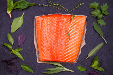 Fresh raw salmon fillet with sage, mint, spinach, cilantro, thyme and rosemary. Healthy food concept on black background. View from above, top studio shot