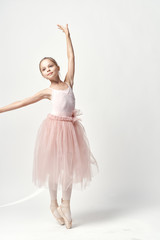 a small ballerina in a light dress and pointe shoes demonstrates her pose on a light background