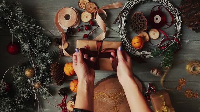 A top-down plan. Fully visible the table with the decorations. Female hands put and finalize Christmas gift wrapped in craftool paper on a wooden table. Bandaging tape and tied in a bow.