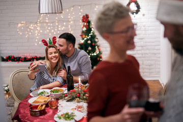 Young cheerful couple taking a selfie at the table while having a party with friends for Christmas holidays.