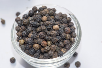 A glass bowl filled with whole black peppercorns on a white surface. Some peppercorns are scattered around the bowl, highlighting their rough texture and rich, dark brown to black color
