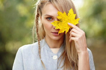 Beautiful young blonde woman covering face with a yellow leaf, autumn concept.