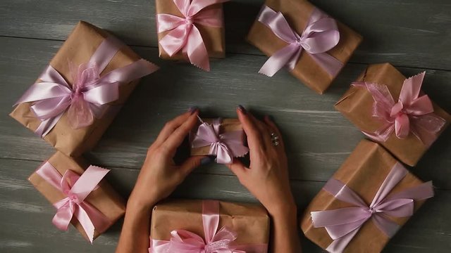 A top-down plan. Fully visible the table with the decorations. Female hands put and finalize Christmas gift wrapped in craftool paper on a wooden table. Bandaging tape and tied in a bow.