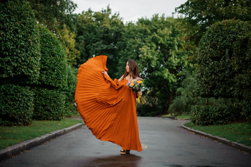 Beautiful woman in long orange dress walking on the wet road