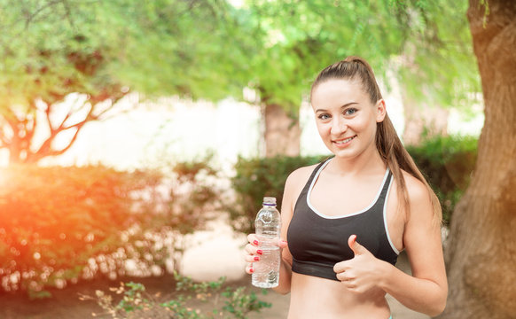 Beautiful, fitness sport girl with ponytail drinks water in the summer green park. Girl drinks water after sport