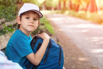 Boy Traveler with a backpack and a cap is sitting by the road and waiting for the bus. Summer vacations outdoor.