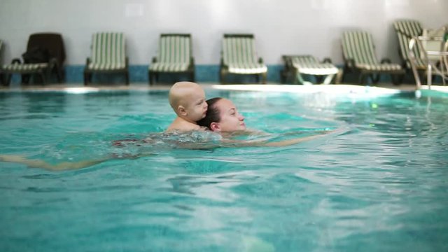 Young mother is swimming in the pool holding her little cute toddler on her back. Adorable little boy is embracing his mother and holding her while swimming in the pool. Piggyback ride in the water