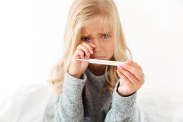 Close-up portrait of sick sad crying little girl holding thermometer, looking at camera