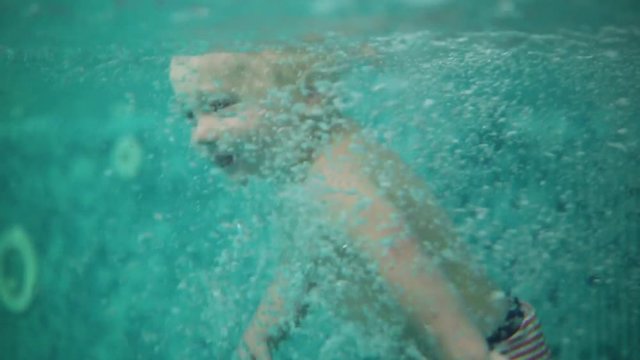 Cute blonde toddler is jumping under the water in the swimming pool and swimming there until his mother is lifting him from the water. An underwater shot