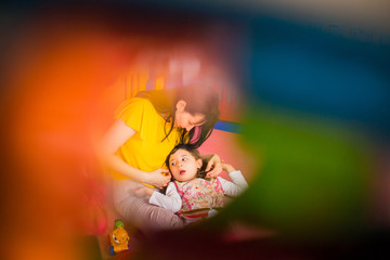 The little beautiful disabled girl lying on lap of young happy mother in the room and colours in the foreground